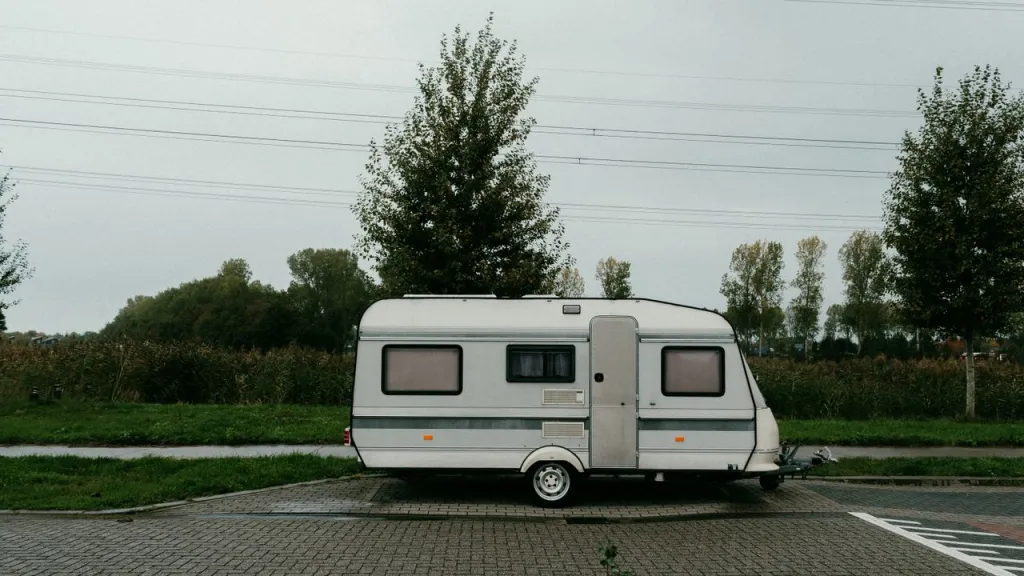 Small white caravan parked on paving beside a green field and trees