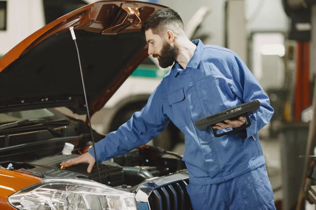 Mechanic in blue overalls holding a tablet while checking the engine bay of an orange car